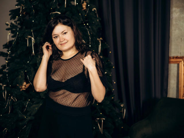 LolaHikls with dark hair, smiling, wearing a sheer black polka dot top, black bra, and black skirt, standing in front of a decorated Christmas tree.