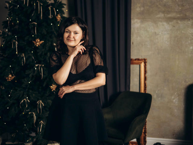 LolaHikls with black mesh top, showing small tits, black skirt, smiling, dark wavy hair, standing near Christmas tree, green chair, and gold frame in dimly lit room.