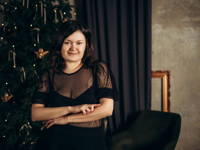 LolaHikls, con un seno di media grandezza, braccia incrociate, sorridente, indossa una maglia nera trasparente, in piedi di fronte a un albero di Natale, una tenda scura e una sedia verde..