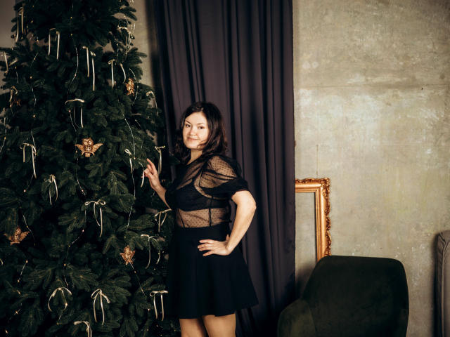 LolaHikls stands next to a Christmas tree, wearing a sheer black top and black skirt, with her hand on her hip, showing a cute smile. She has small tits.