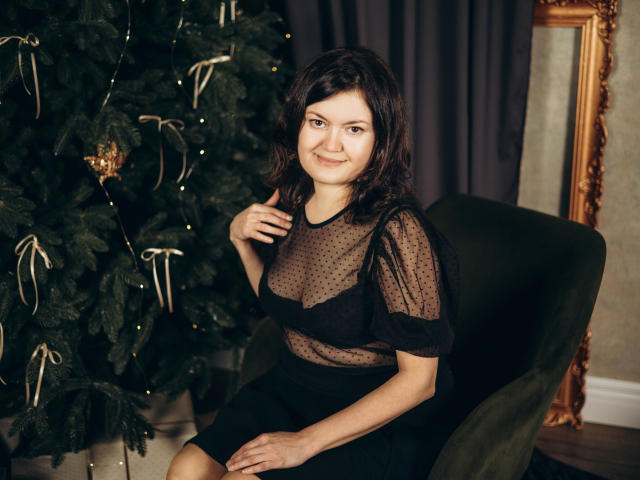 LolaHikls con un seno di media grandezza, che indossa una maglietta nera trasparente a pois, seduta su una sedia verde, sorride alla fotocamera, accanto a un albero di Natale decorato..