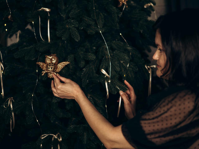 LolaHikls, con i capelli castani, indossa una trasparente maglia nera e tiene in mano un ornamento a forma di stella dorata e scintillante su un albero di Natale verde scuro, guardando di lato con un sorriso appena accennato..