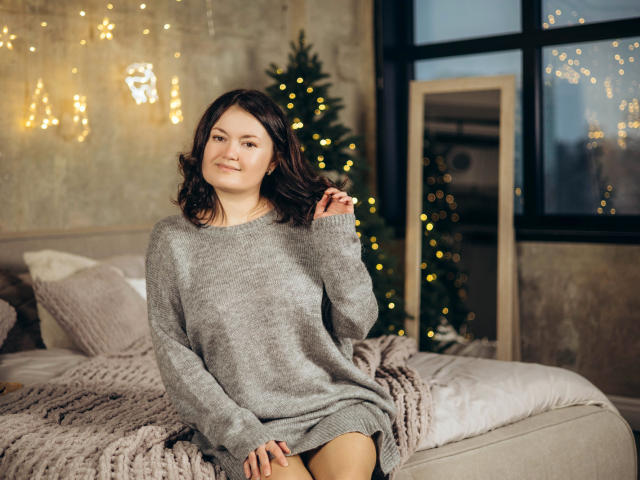 LolaHikls with wavy brown hair, smiling, wearing a gray knit sweater, sitting on a bed with a knitted blanket, hands near face, Christmas tree with lights in background.