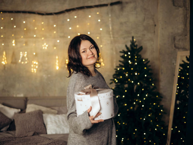 Lola, con capelli castani lunghi fino alle spalle, indossa un maglione grigio, tiene in mano una scatola regalo bianca con un nastro dorato, sorride e si trova di fronte a un albero di Natale con luci gialle..