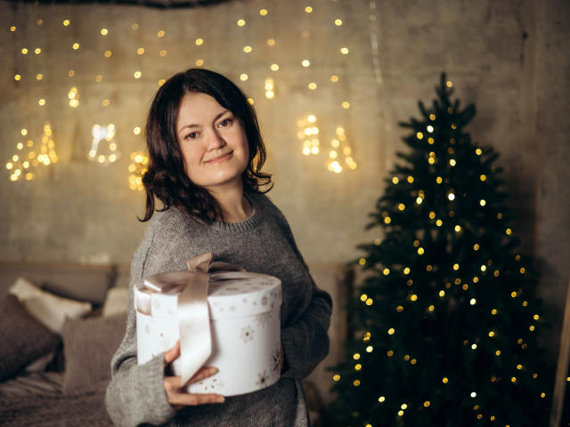 Lola, con capelli castano ondulati, indossa un maglione grigio, tiene in mano una scatola regalo bianca con un nastro rosa, sorride alla telecamera e si trova accanto a un albero di Natale decorato con luci gialle..