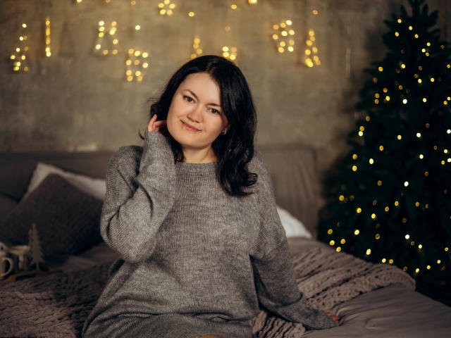 LolaHikls, with wavy black hair, smiles at the camera wearing a gray, long-sleeve sweater, sitting on a bed with a Christmas tree and warm lights in the background.