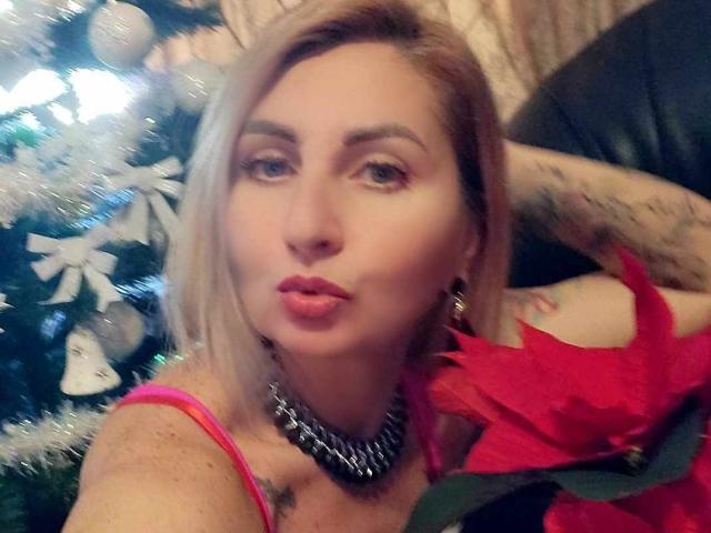 ChaudeEvely with blonde hair, pouty lips, wearing a pink top, black necklace, and red poinsettia, has tattoos on her arm, and a Christmas tree in the background.