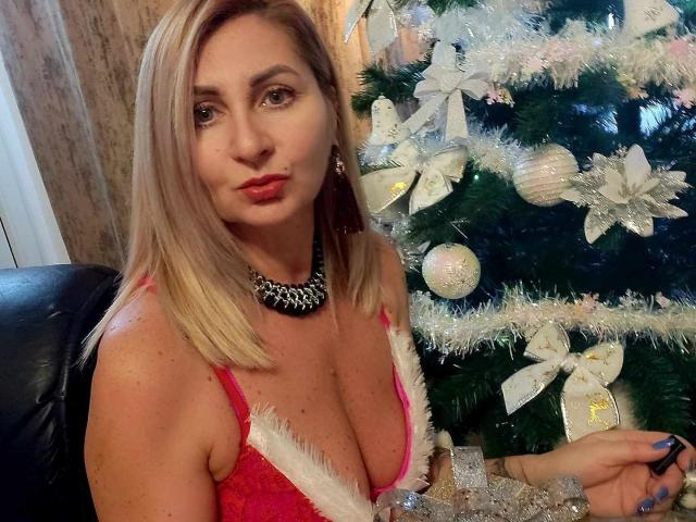 ChaudeEvely with blonde hair, red lingerie, and white fur trim, showing her large tits, wearing a black necklace, sitting next to a decorated Christmas tree.