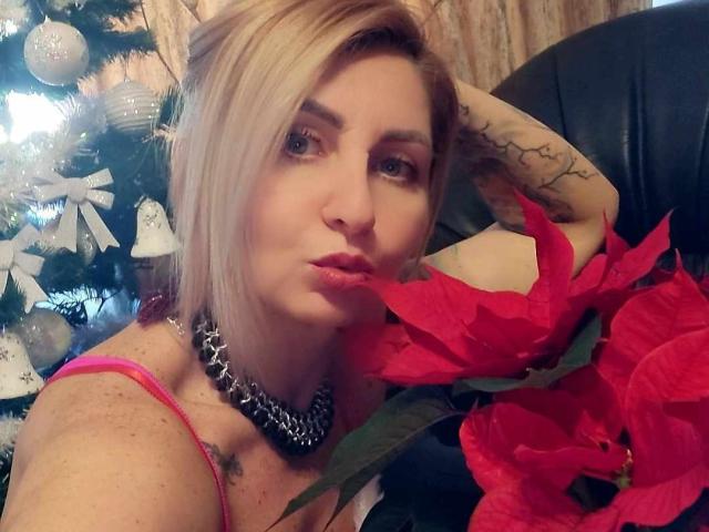 ChaudeEvely with blonde hair, pink top, chain necklace, pouty lips, tattooed arm, holding red poinsettia, Christmas tree with white ornaments in background.