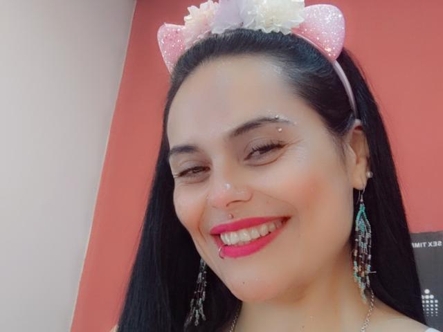CharlotteCrofort with black hair, pink glitter cat ears, sparkling nose, red lipstick, tongue piercing, and colorful earrings, smiling at the camera.