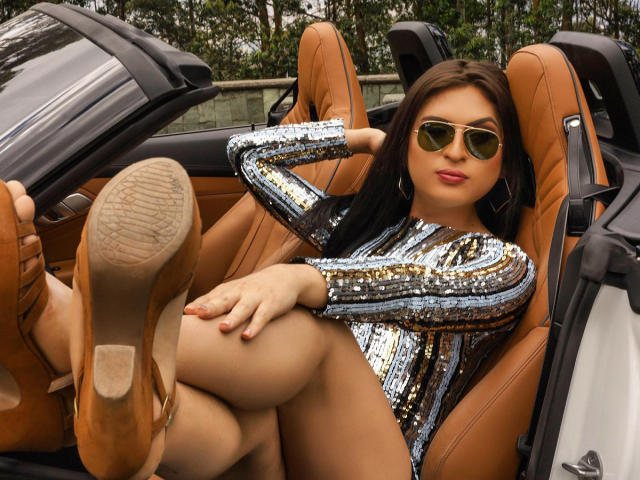 SthefaniaM with brown hair, wearing gold sunglasses, a shiny silver sequin dress, and tan heels, lounging in a brown leather car seat, legs up, showing her bare ass.