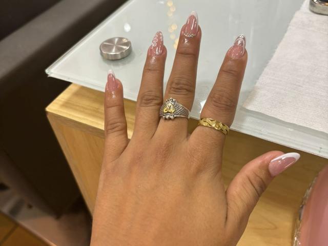A mão de EimmyPark, com unhas rosadas e brilhantes, usando um anel de ouro com um diamante e uma gema amarela, em um balcão de vidro com um suporte de anel de metal..
