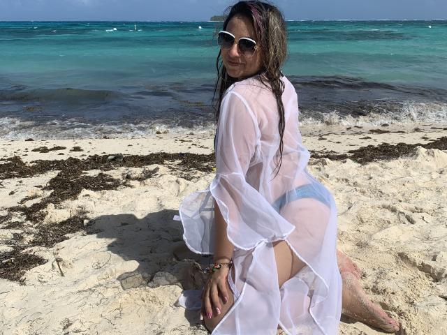 AmandaLeon on a beach, wearing a sheer white robe, showing her blue panties and ass, with wet brown hair and white sunglasses, kneeling on sand.