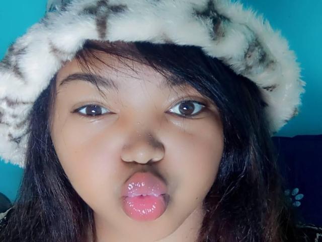 AnnayahOne with puffy pink lips, wearing a fluffy white and black hat, has brown eyes and black hair, looking slightly up with a playful expression.