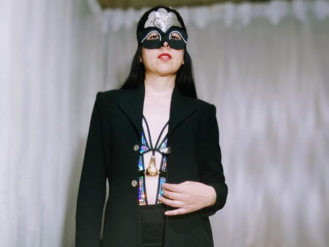RossanaHairy wearing a black mask with a silver gem, black blazer, and multicolored necklace, standing with hand on belly, white background. Her tits are small, she's cute.