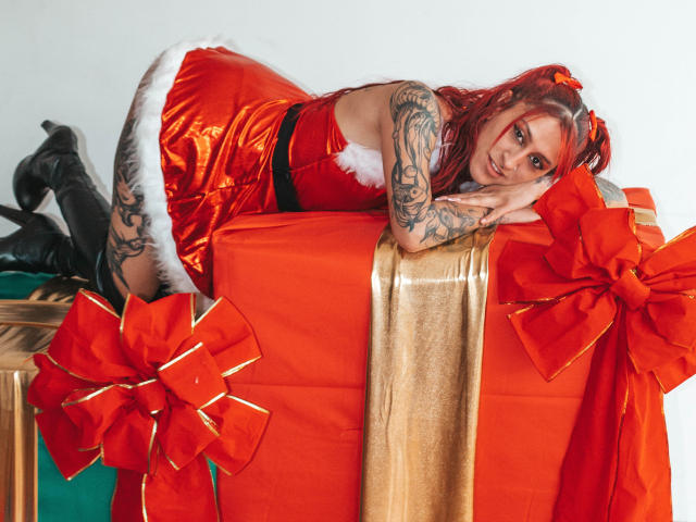 SofiFw with red hair, tattoos, and a sexy Santa dress, leaning on a giant orange gift with red bows, black thigh-highs, and a white fur trim.
