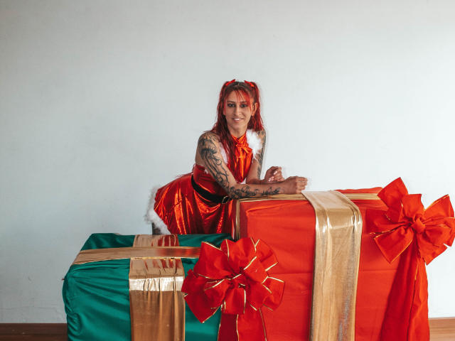 SofiFw with red pigtails, tattooed arm, smiling, wearing shiny red dress, leaning on large gift wrapped in green, red, and gold with red bows.