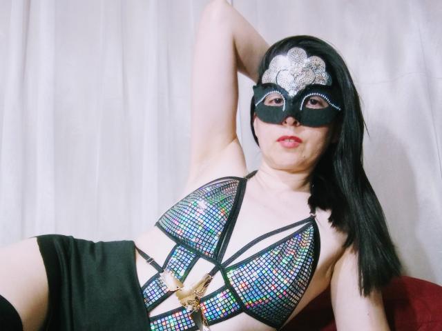 RossanaHairy with black mask, wearing iridescent bra and black shorts, has long black hair, fair skin, and red lips, showing her tits and ass in a sexy pose.