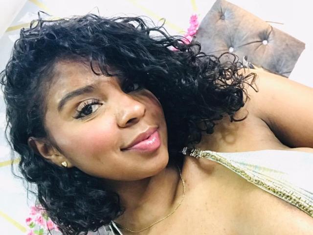 LisaMendez with curly black hair, brown skin, and pink lips, wearing a gold necklace and a strapless top, smiling at the camera. Her small tits are partially visible.