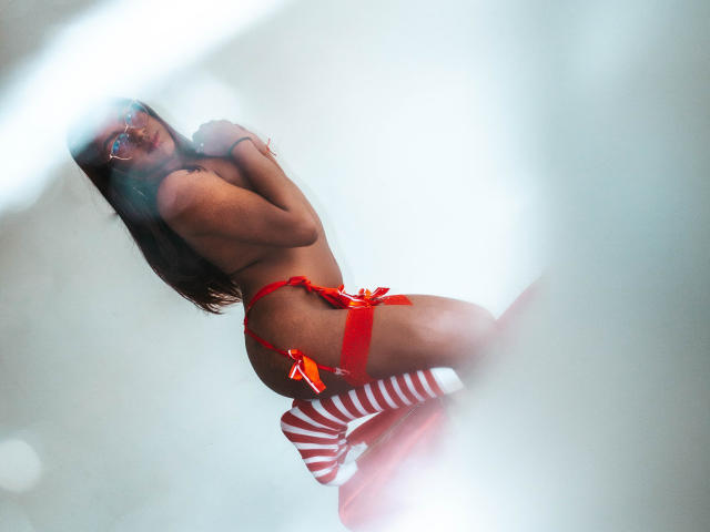 KimmySolange in red lingerie with red ribbon, striped socks, and glasses, showing her ass and tits, kneeling, looking back, in a blurred, soft-focus shot.