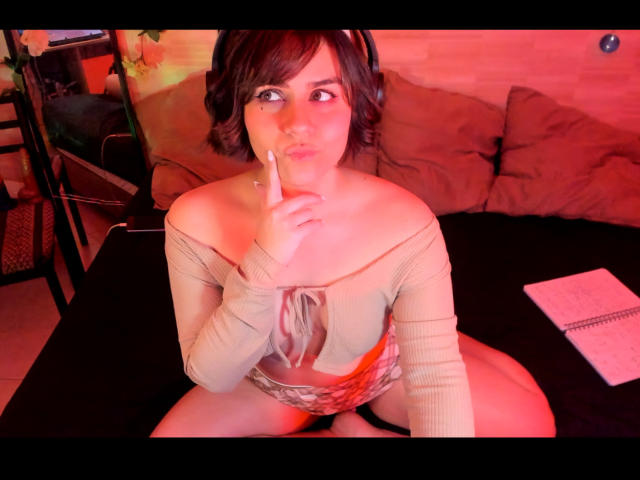 IsabelaCooper with short brown hair, wearing a beige off-shoulder top, index finger to lips, sitting on black table, red lighting, headphones on, notebook with pen on right.