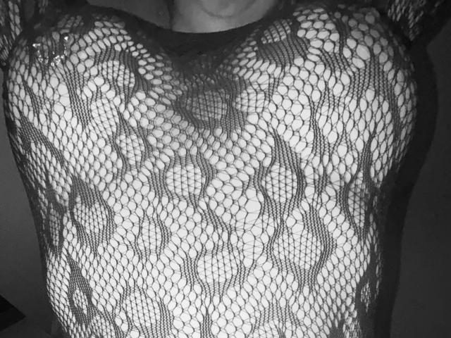 DarkWolfCouple's chest in a black fishnet lace top, showing their boobs through the net pattern. The lace is tight, revealing their nipples and areolas underneath. Black and white.