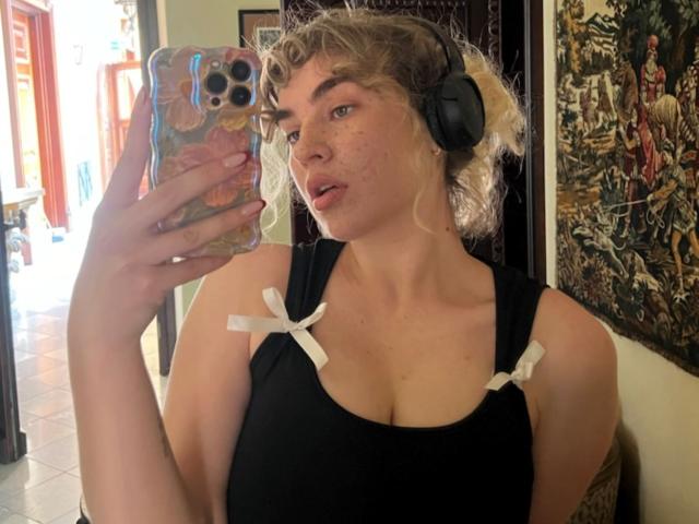 Marilyn, speels met krullend blond haar, een zwart tanktop met witte linten, een koptelefoon, die een telefoon vasthoudt en zichzelf in de spiegel fotografeert, waarbij haar borsten zichtbaar zijn. Haar telefoonhoesje is kleurrijk en heeft een patroon..