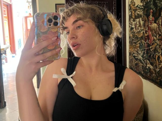 Marilyn: Capelli biondi ricci, canotta nera con fiocchi bianchi, cuffie, tiene in mano un telefono con una custodia rosa, guarda allo specchio, mostrando il seno, le lentiggini e la bocca leggermente aperta..