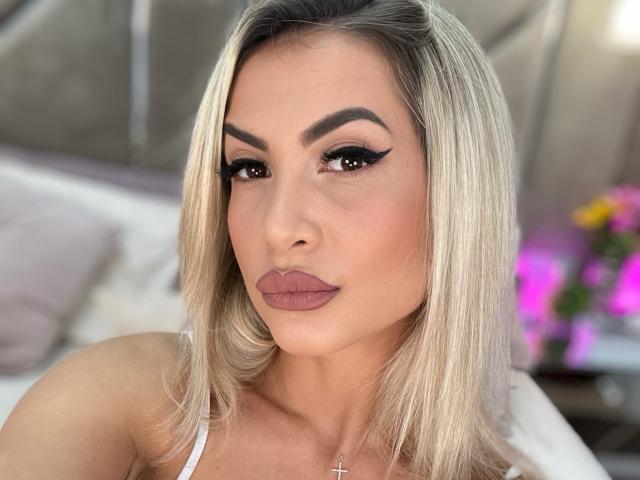Lellie has blonde hair, thick eyebrows, black eyeliner wings, and brown lipstick. She's wearing a white top and a cross necklace. Her boobs are slightly visible.