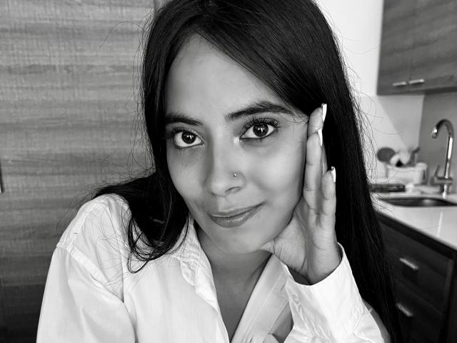 TamaraPeach with long dark hair, nose piercing, white shirt, and hand on cheek, smiling in black-and-white photo. Her eyes are large and she has a cute, relaxed expression.