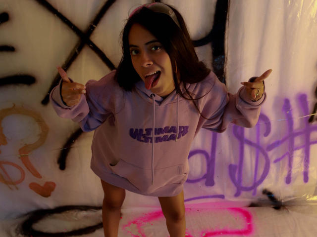 TamaraPeach, con cabello oscuro, sacando la lengua, vistiendo una sudadera morada con la inscripción "UASTART", señalando con los dedos, de pie con las piernas separadas, en un espacio tenuemente iluminado y cubierto de grafitis..