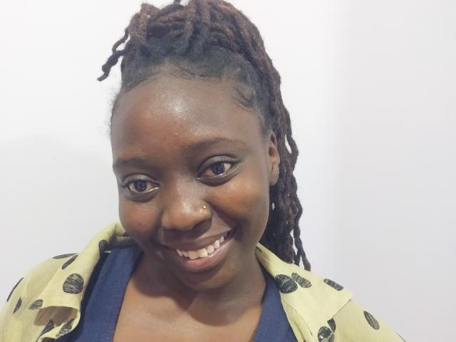 Lushmourine has dark brown skin, small nose, smiling with teeth showing, brown eyes, and dreadlocks. She wears a yellow jacket with black patterns over a blue shirt.