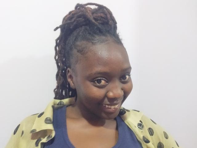 Lushmourine with dark brown skin, curly red-brown dreadlocks, yellow jacket with black polka dots, blue shirt, smiling slightly, looking to the side.