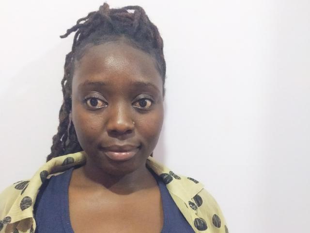 Lushmourine has dark brown skin, small nose, and is smiling with her lips closed. She's wearing a beige shirt with black patterns over a blue top, and has braided hair.