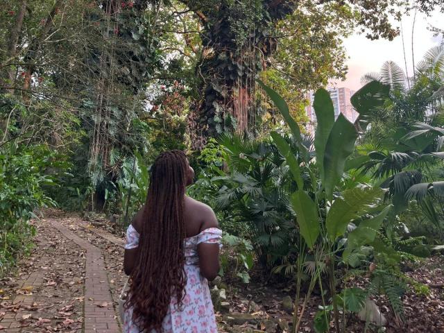 AkaneCox with long, dark brown, curly hair stands on a brick path, wearing an off-shoulder white dress, surrounded by lush greenery and large banana leaves.