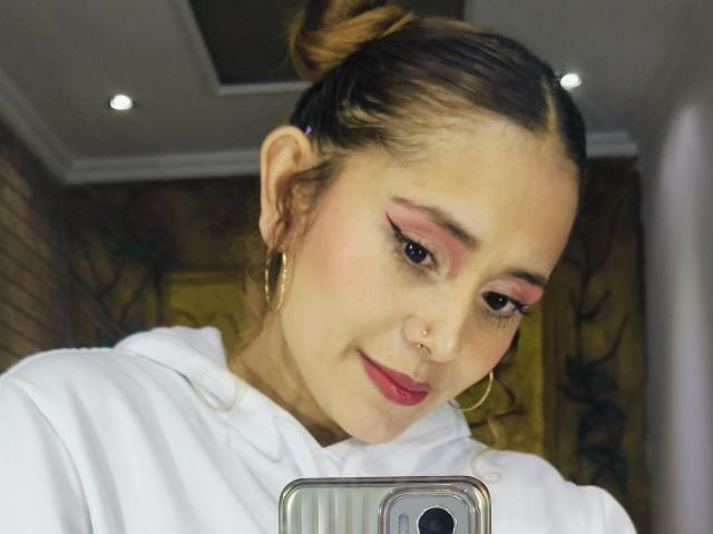 EliBaker mit braunen Haaren zu einem Dutt gebunden, pinkem Eyeliner, rotem Lippenstift, Nasenring, goldenen Creolen, weißer Kapuzenjacke, die ein Selfie mit einem Handy vor einem Spiegel macht..