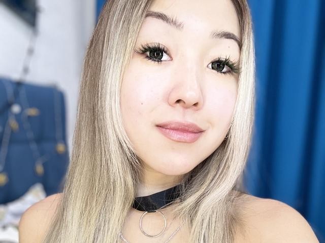 RenyLime has blonde hair, green eyes, and a cute face with pink lips. She's wearing a black choker with a ring, and has long, thick, black eyelashes.
