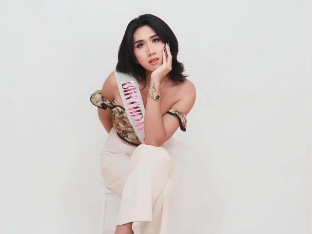 NathalieLore with black shoulder-length hair, wearing an off-shoulder floral dress, sitting with one knee up, hand on cheek, looking at camera, white background, showing her tits.