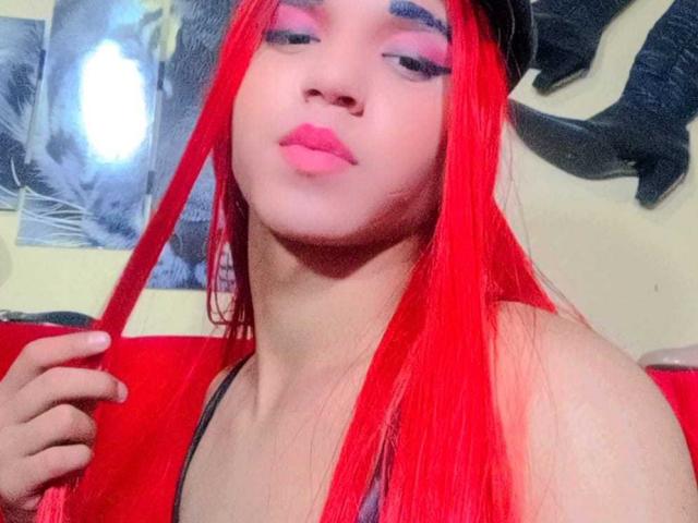 AnggieTs has bright red hair, pink eyeliner, and pink lipstick, holding her hair with a black hat on. She's wearing a red top, showing her tits.