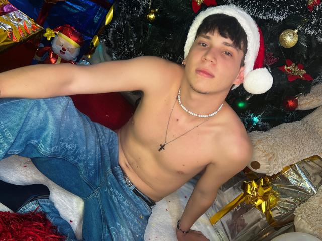 JonathanMoreau, shirtless with a Santa hat, wearing blue jeans and a pearl necklace, sitting in front of a Christmas tree with gifts and a stuffed toy.