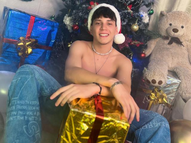 JonathanMoreau, shirtless, wearing a Santa hat, blue jeans, necklace, smiling, sitting in front of a Christmas tree with gifts, and a large teddy bear, looking cute and happy.