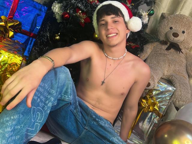 Shirtless JonathanMoreau with a Santa hat, smiling, wearing blue jeans and a necklace, sitting among Christmas gifts and a giant teddy bear. He has a cute, slim, attractive build.