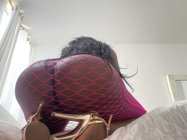 DiosaEbano's ass in pink fishnet, kneeling on a bed, showing big round butt, wearing gold high heels, black hair, white walls in the background, taken from a low angle.