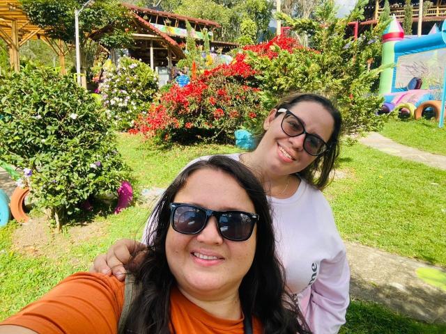 CarlaNaughty with black hair, orange shirt, and sunglasses in front, CarlaNaughty with brown hair, glasses, and white shirt smiling behind, in a sunny garden with colorful plants.