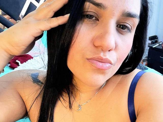 VivianaThompson has black hair, a tattoo on her right shoulder, and is pouting her pink glossy lips while touching her head with a hand, wearing a blue strap top.