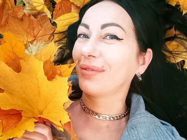 MirandaHott69 sdraiata su un letto di foglie autunnali gialle e arancioni, indossa una collana d'argento e una giacca di jeans blu, con eyeliner nero e un leggero sorriso..