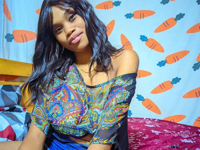 Tiah with dark wavy hair, small tits, wearing a colorful, paisley-patterned, off-shoulder top, sitting on a bed with red and pink patterned sheets.
