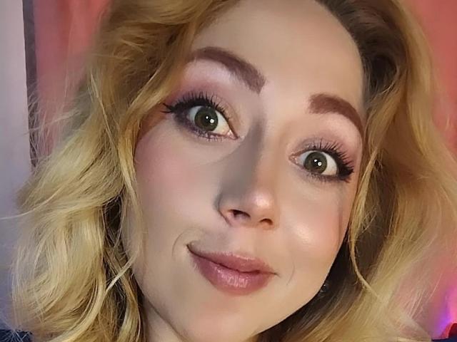 DouceKhrystine has blonde wavy hair, green eyes, thick eyebrows, and pink eyeshadow with long eyelashes, smiling with glossy pink lips. Her skin is fair and she's looking at the camera.
