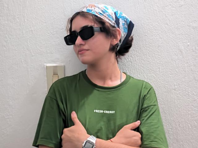 BrianaSoica with brown hair, wearing a blue patterned headscarf, black sunglasses, green shirt, silver watch, and crossed arms, giving a thumbs-up, with a neutral expression.