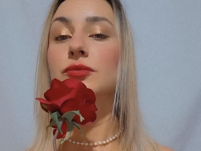 CherryDouce, con una rosa roja en la boca, lleva un collar de perlas, tiene sombra de ojos dorada brillante y lápiz labial rojo, y tiene el cabello rubio. Sus glúteos no son visibles..